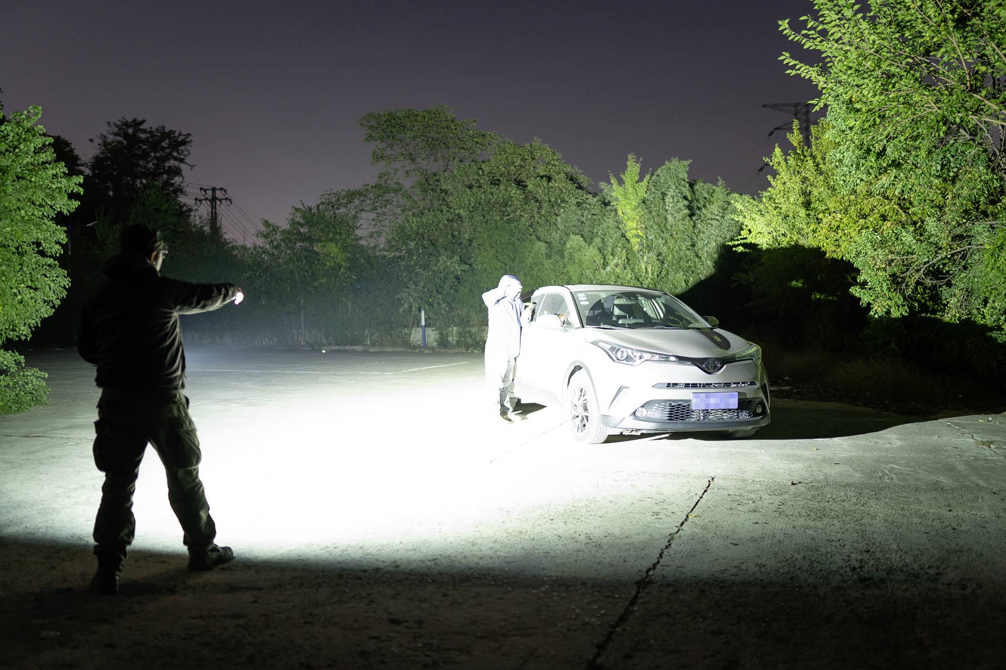 Zwei Personen stehen nachts neben einem beleuchteten Auto auf einer Straße, umgeben von Bäumen im Hintergrund.
