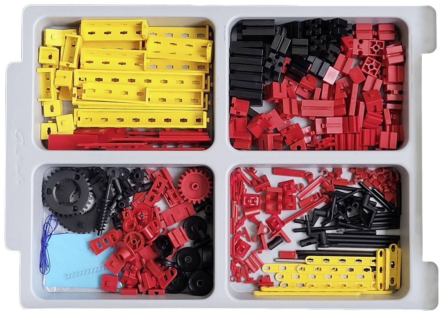 Ein geöffneter Kunststoffkasten enthält sortierte Baukastenteile: oben links gelbe Balken, oben rechts rote und schwarze Teile, unten rote Zahnräder und schwarze Verbindungsstücke.