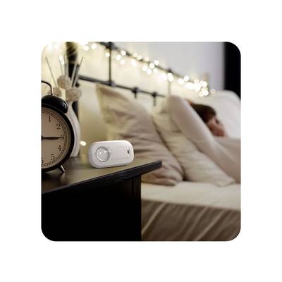Eine Person liegt entspannt auf einem Bett im Hintergrund, während im Vordergrund ein Wecker und ein elektronisches Gerät auf einem Tisch stehen.