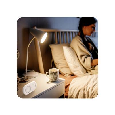 Frau sitzt im Bett mit Laptop im Hintergrund; im Vordergrund leuchtet eine Lampe neben einem smarten Lichtschalter auf dem Nachttisch.