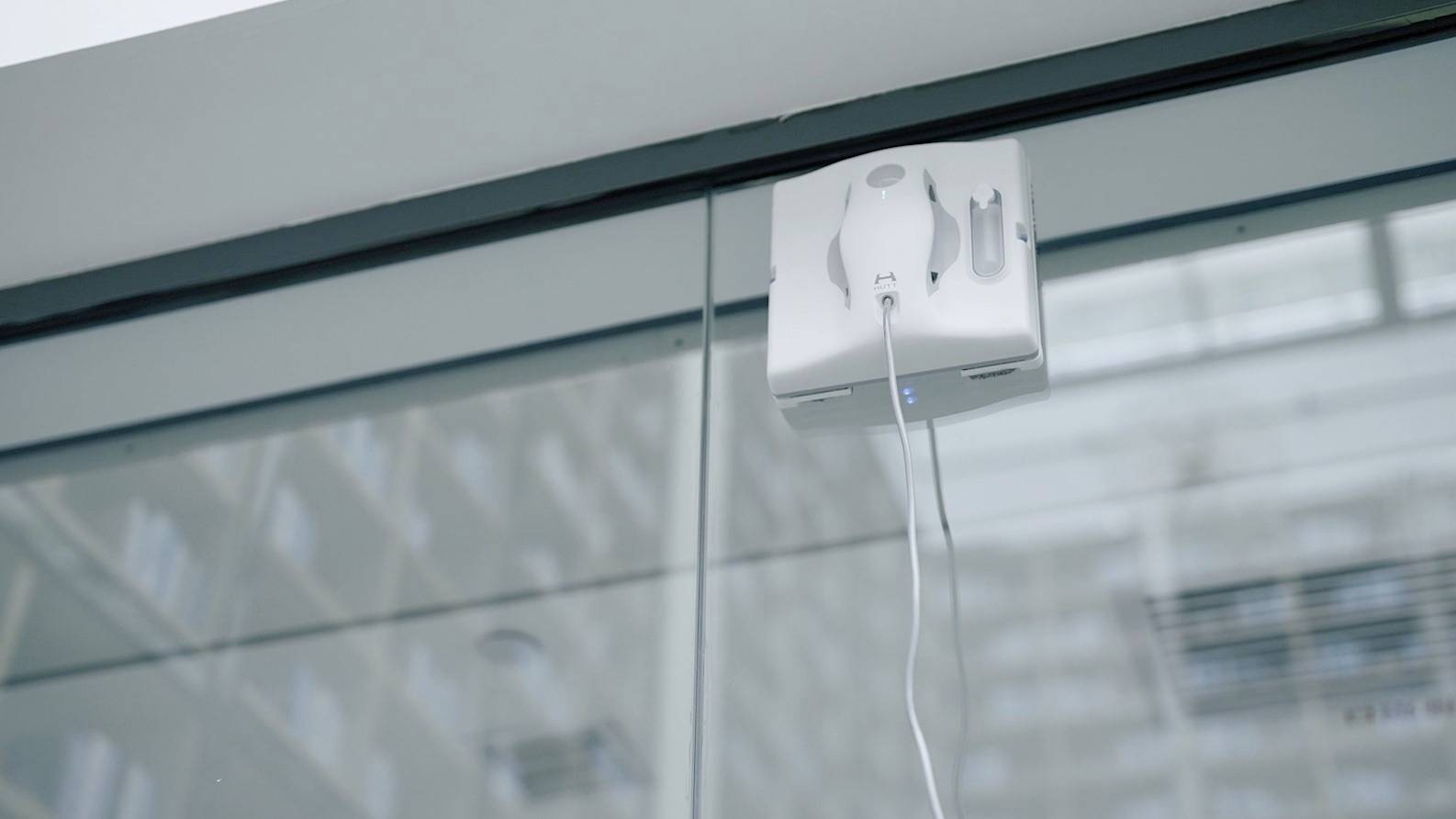 Ein Fenstersaugroboter ist an einer großen Glasfläche befestigt und wird von einem Kabel mit Strom versorgt, während er arbeitet.