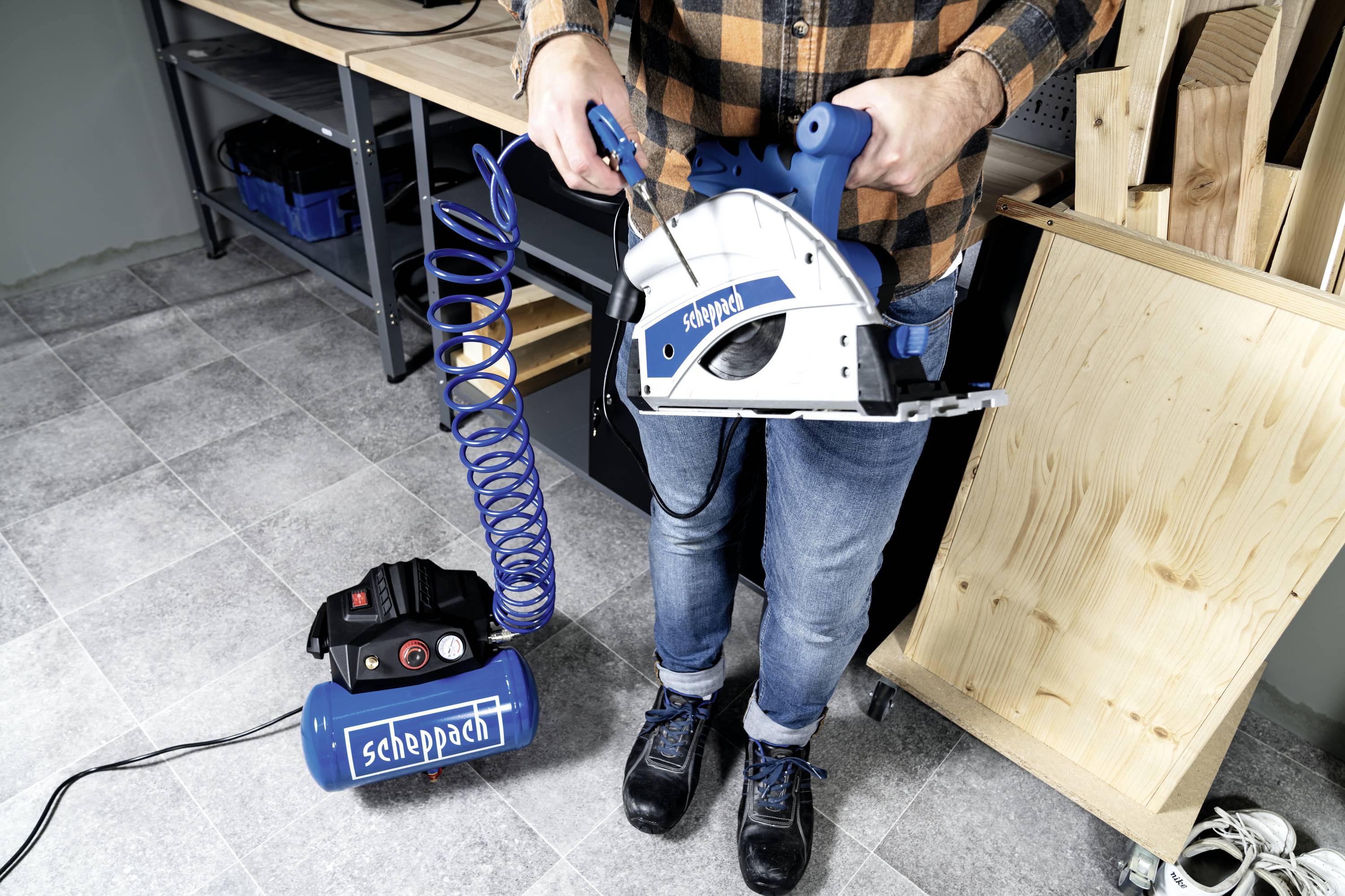 Person hält eine blaue Druckluftsäge 'Scheppach' in der Hand, mit einem Kompressor verbunden. Hintergrund zeigt Werkstattumgebung.