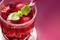 Ein Glas roter Fruchtsaft mit Eiswürfeln, Minzblättern und einem gestreiften Strohhalm vor einem pinken Hintergrund.