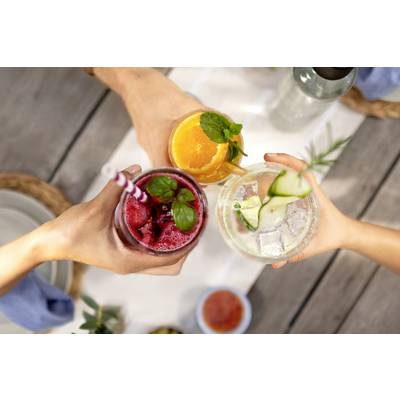 Drei Personen stoßen mit farbigen Cocktails an, die mit Minzblättern, Orangenscheiben und Gurkenscheiben dekoriert sind.