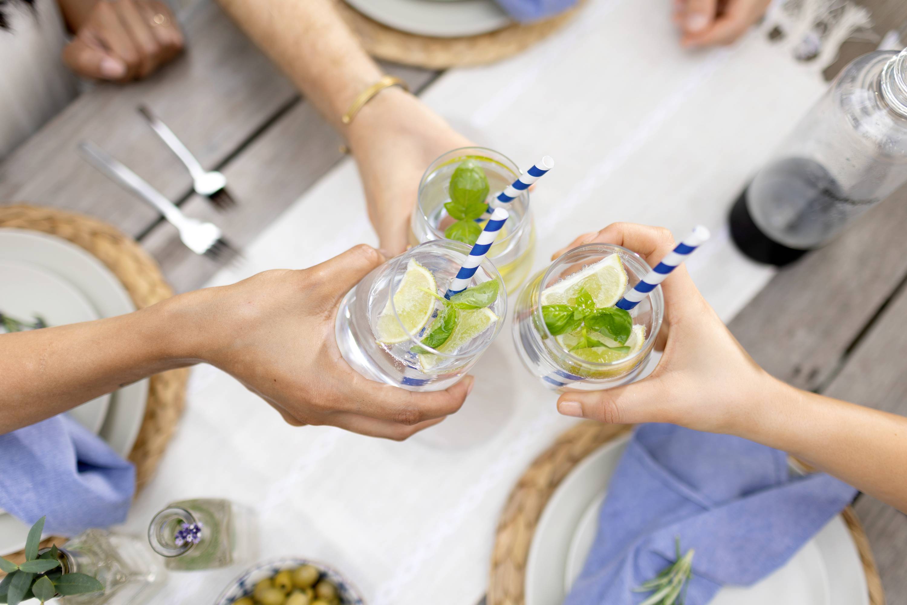 Drei Personen stoßen mit Gläsern gefüllt mit Zitronenlimonade an. Die Szene findet an einem gedeckten Esstisch im Freien statt.