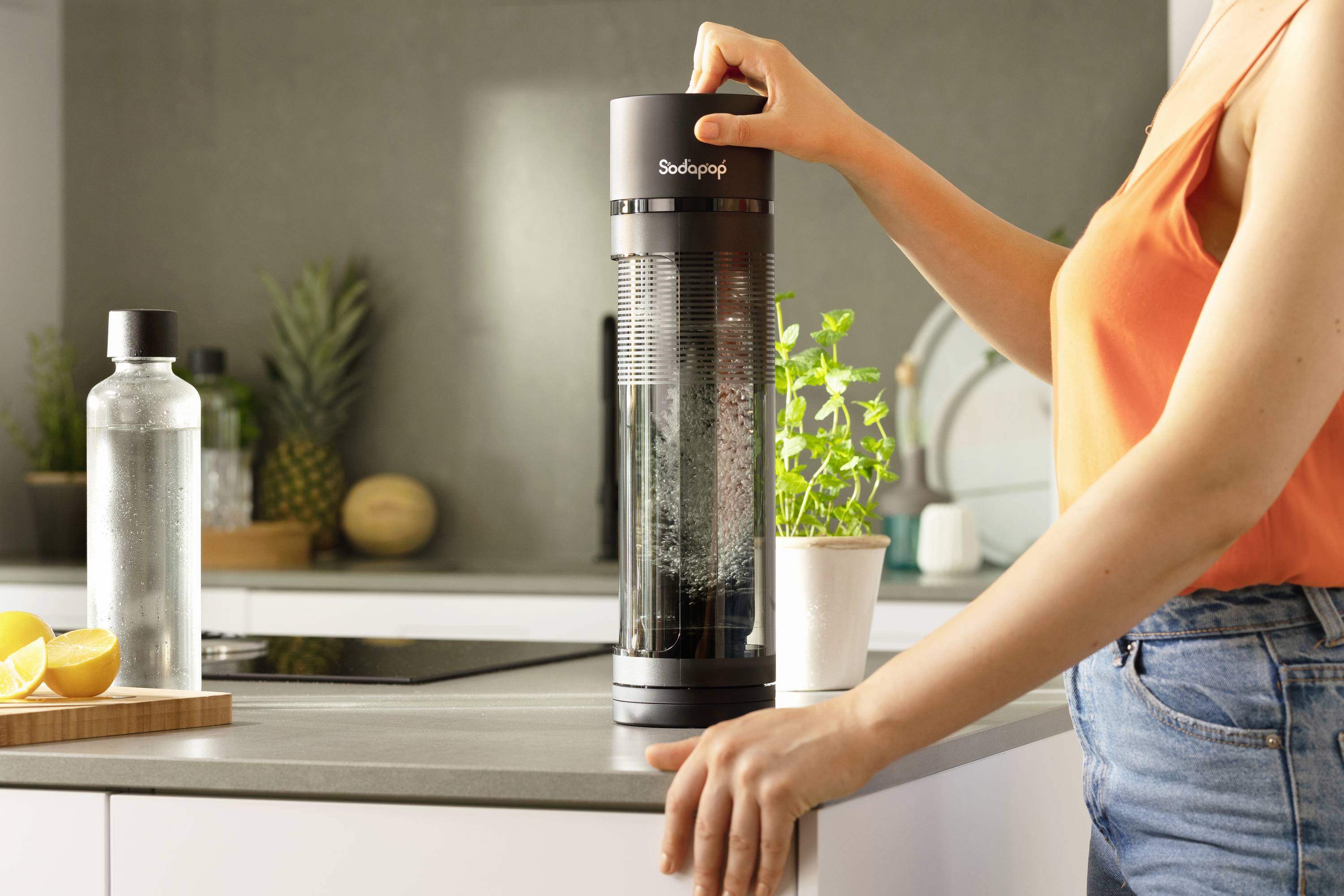 Person benutzt einen Wassersprudler auf einer Küchenarbeitsplatte. Im Hintergrund sind Küchenutensilien und eine Pflanze zu sehen.