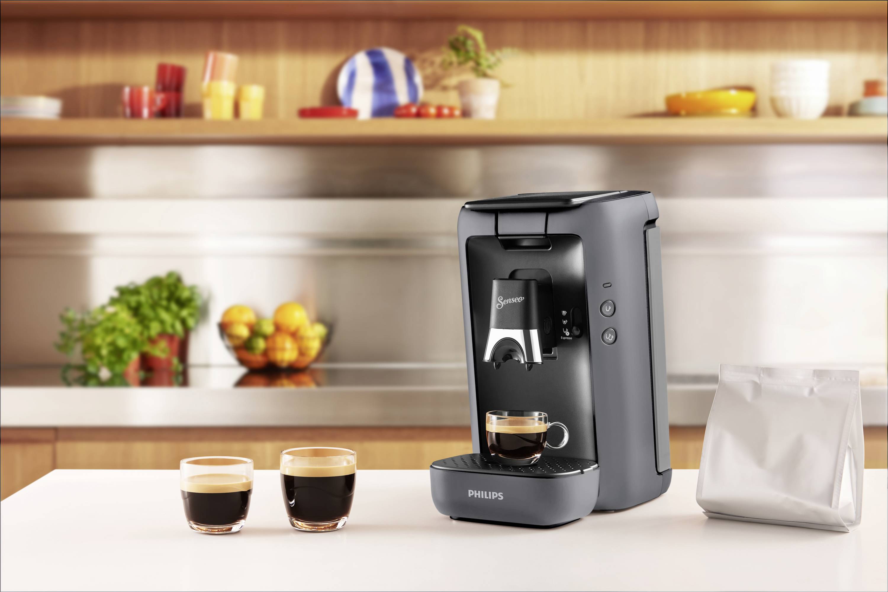 Eine graue Kaffeemaschine steht auf einer Küchenarbeitsplatte mit zwei gefüllten Kaffeetassen und einem Beutel in der Nähe. Im Hintergrund sind Regale mit Geschirr und Obst.