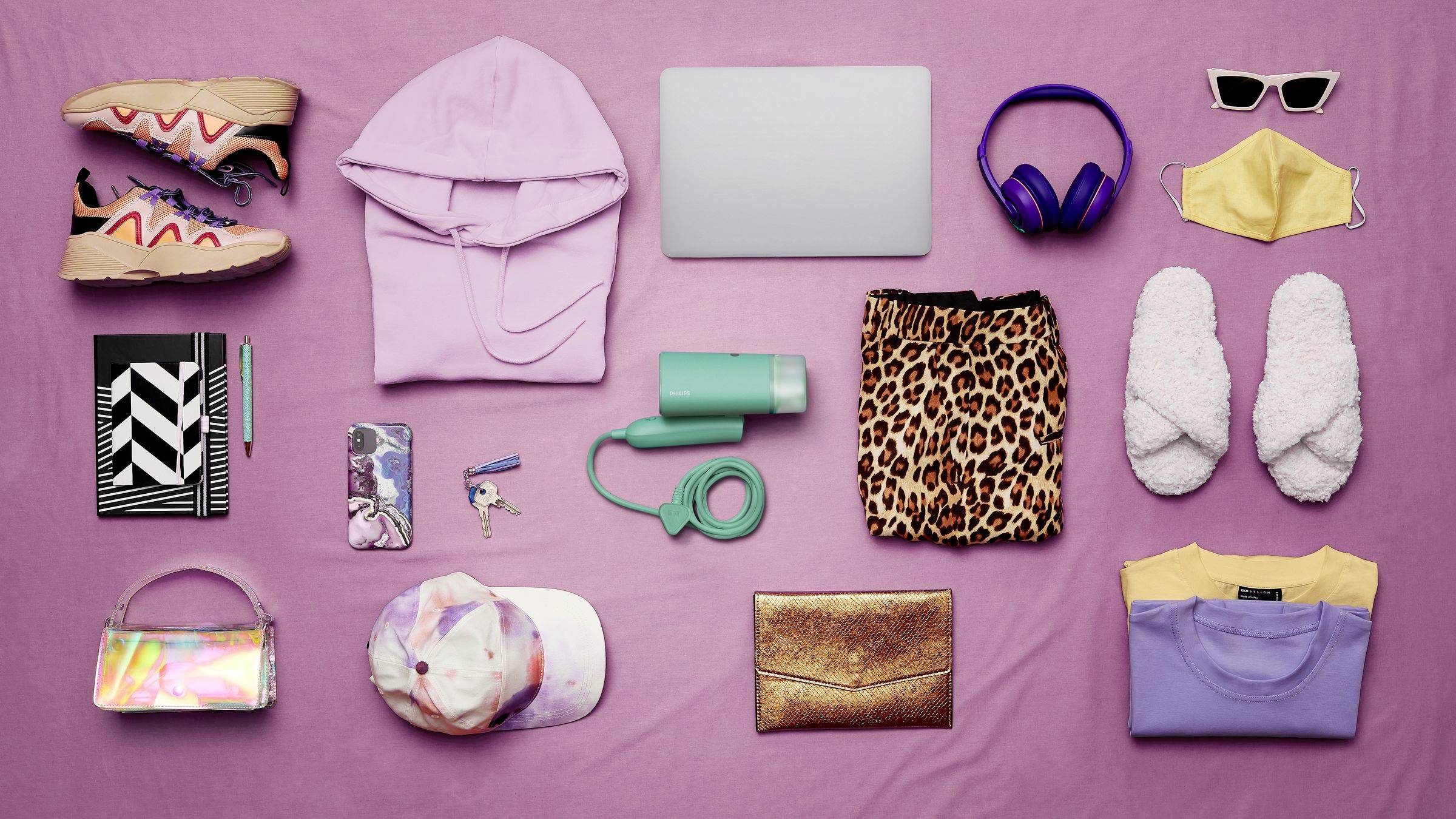 Verschiedene Gegenstände auf einem rosa Hintergrund, darunter Schuhe, Kleidung, Laptop, Notizbuch, Kopfhörer, und Accessoires.