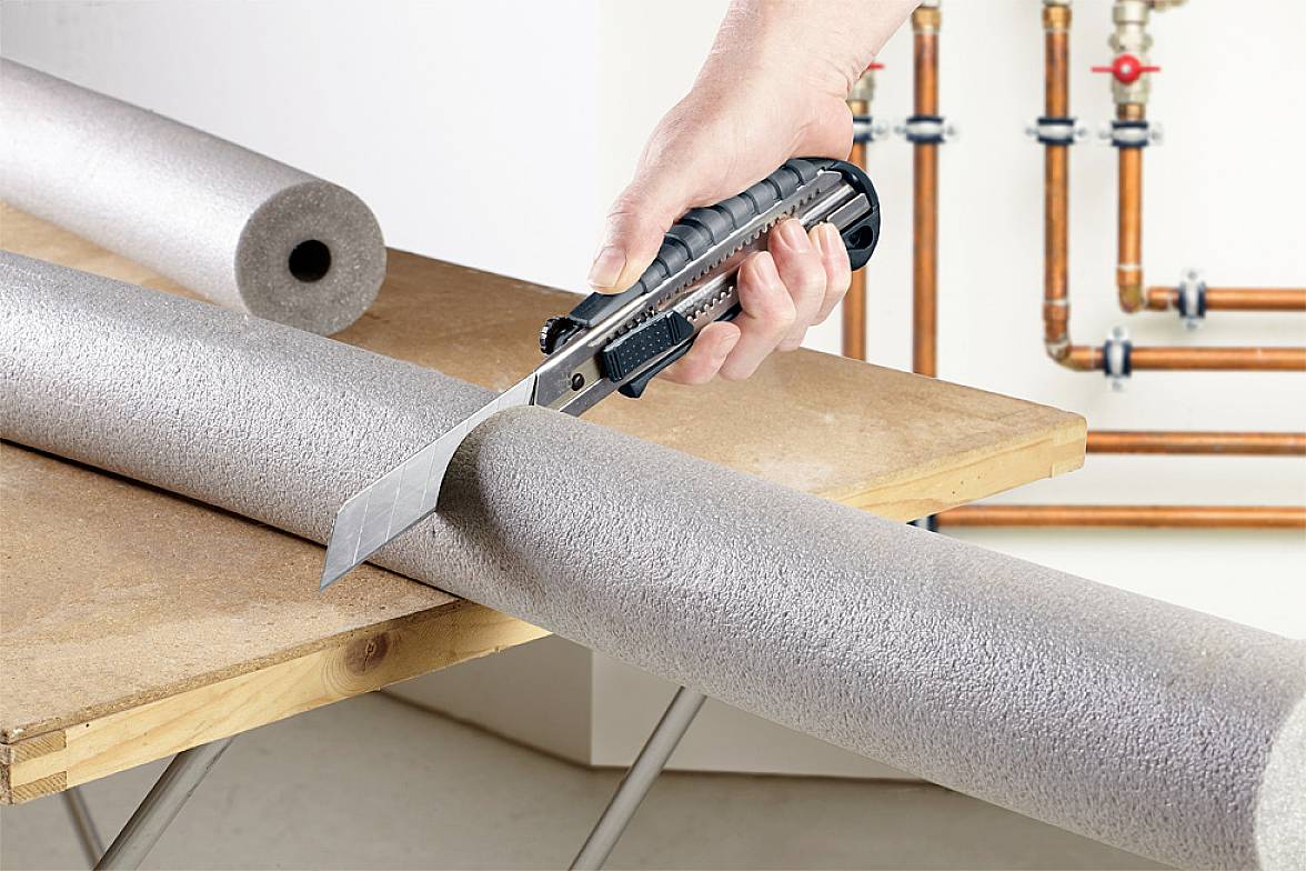 Eine Hand schneidet mit einem Cutter-Messer eine graue Rohrisolierung auf einem Tisch. Im Hintergrund sind Kupferrohre sichtbar.