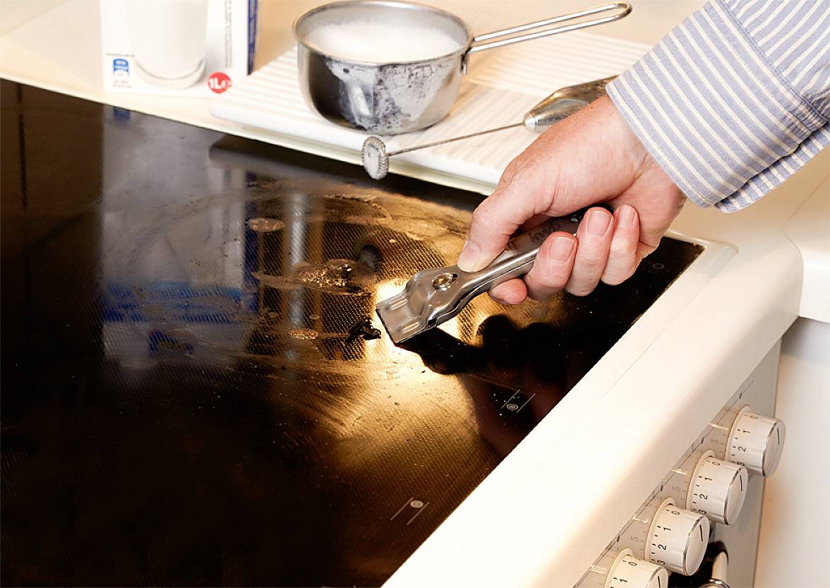 Eine Person reinigt ein Kochfeld mit einem Schaber. Im Hintergrund ist ein Topf mit warmem Seifenwasser. Die Szene zeigt Küchenreinigung.
