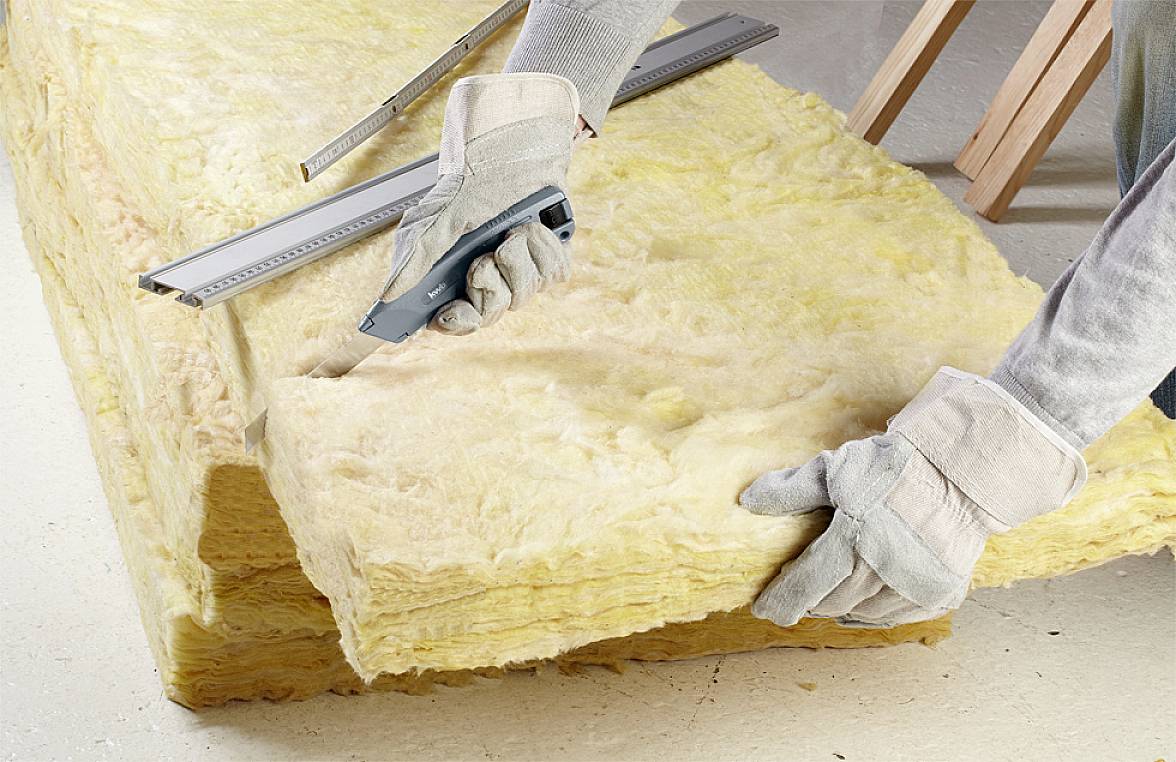 Eine Person mit Handschuhen schneidet ein Stück gelbe Isolierung mit einem Messer und einem Lineal, wahrscheinlich im Bauumfeld.