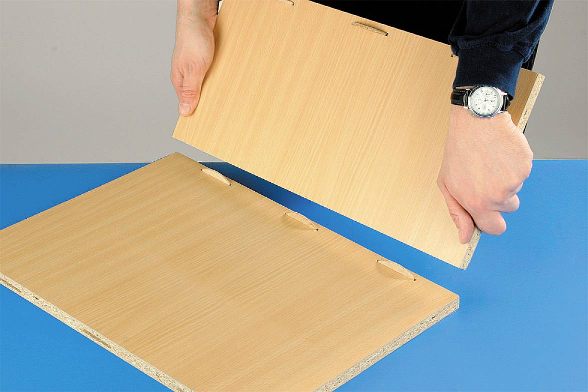Person hält zwei Holzpaneele, die verbunden werden. Eine Hand zeigt eine Uhr. Die Paneele sind auf einem blauen Tisch platziert.