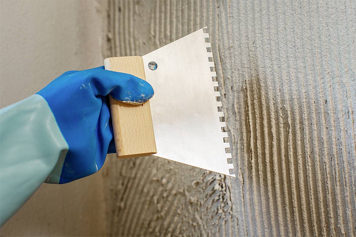 Eine Person mit blauen Handschuhen trägt mit einer gezahnten Kelle eine gleichmäßige Schicht Kleber auf eine Wand auf.