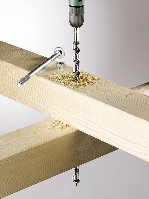 Eine grüne Bohrmaschine bohrt ein Loch in einen Holzbalken. Holzspäne liegen um das Bohrloch herum. Ein Schraubenschlüssel liegt in der Nähe.