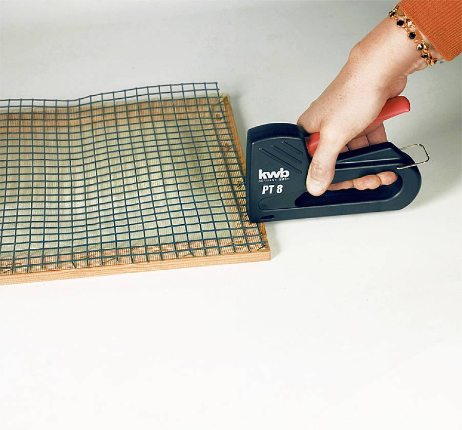 Eine Hand hält einen Tacker und befestigt ein Drahtgitter auf einer Holzplatte.