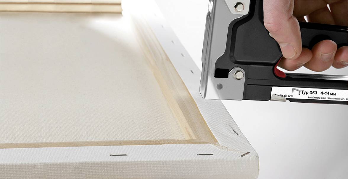 Eine Hand benutzt einen Tacker, um eine Leinwand mit Holzrahmen zu befestigen. Der Tacker fixiert den Stoff entlang des Rahmens.
