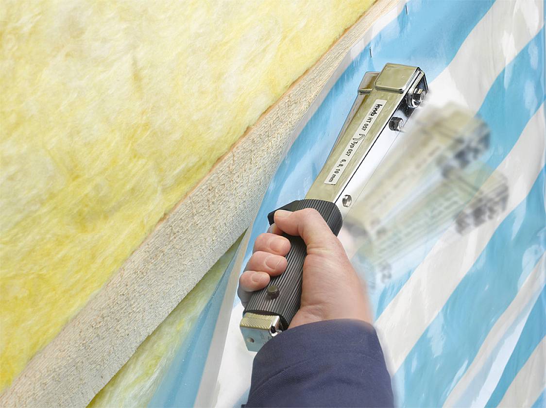 'Hand hält Klammergerät' befestigt Isoliermaterial an blau-weiß gestreifter Oberfläche. Hauptaktion ist Isolierung anbringen.