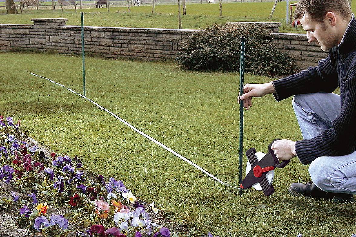Ein Mann misst ein Stück Rasen neben einem Blumenbeet, indem er ein Maßband entlang des Bodens zieht. Im Hintergrund eine Steinmauer.