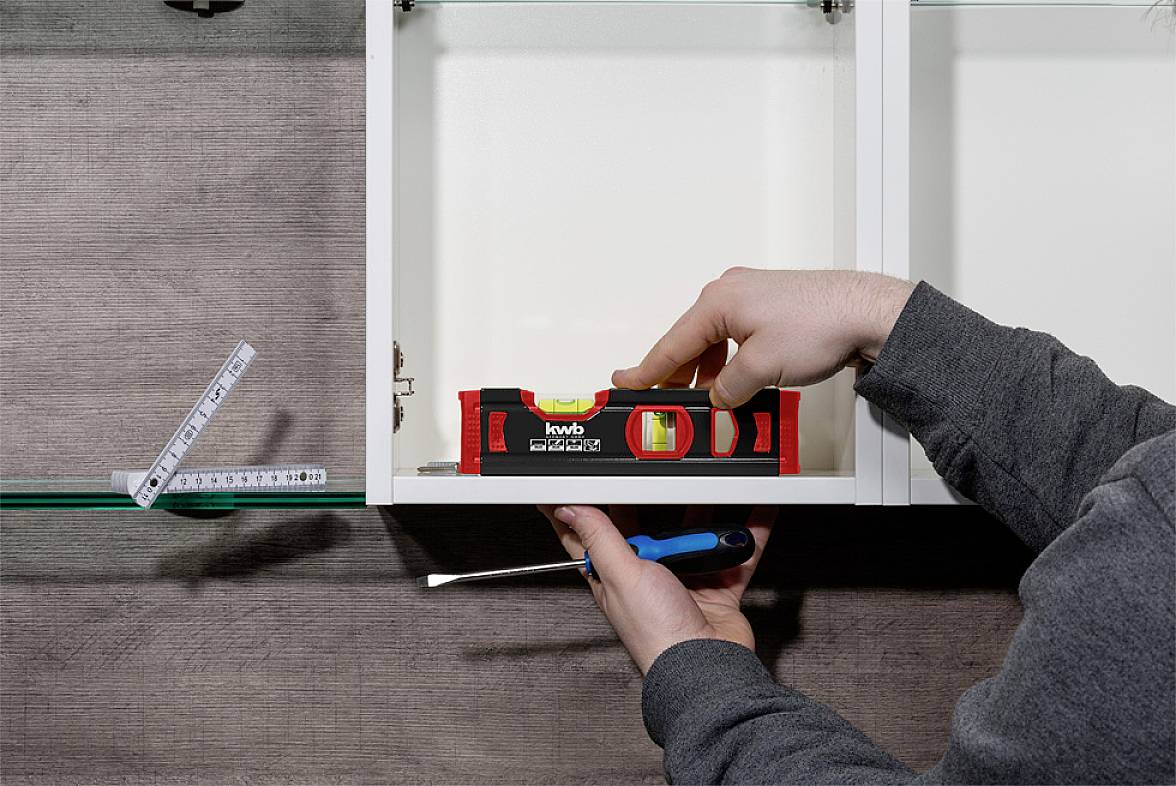 Person misst einen Schrank mit einer Wasserwaage und einem Schraubendreher. Ein Maßband liegt in der Nähe.