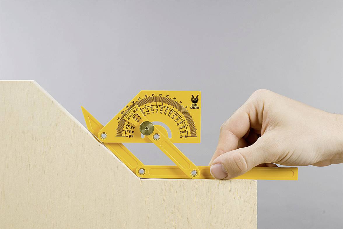 Eine Hand hält einen gelben Winkelmesser an einem Holzstück, um den Winkel einer Kante zu messen. Der Winkelmesser zeigt ca. 45 Grad an.