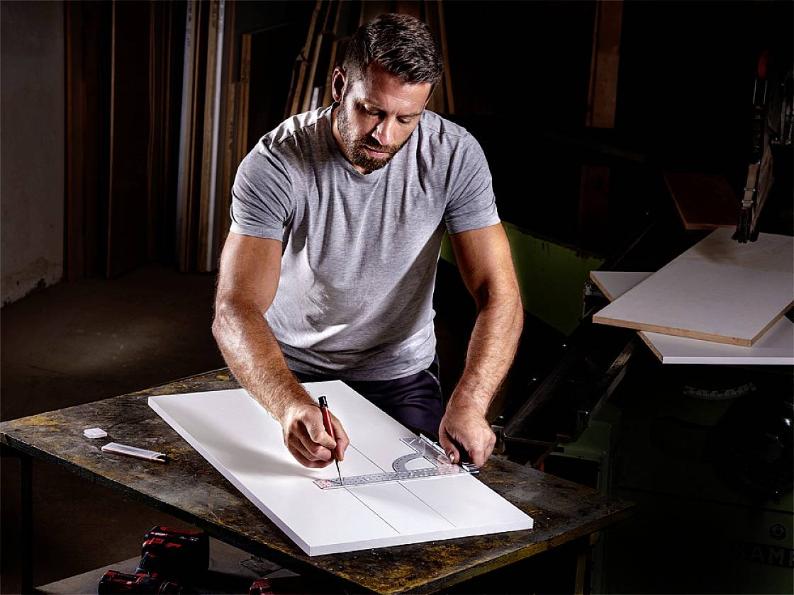 Ein Mann arbeitet konzentriert an einem Tisch mit Holzplatten, zeichnet mit einem Bleistift auf eine der Platten.