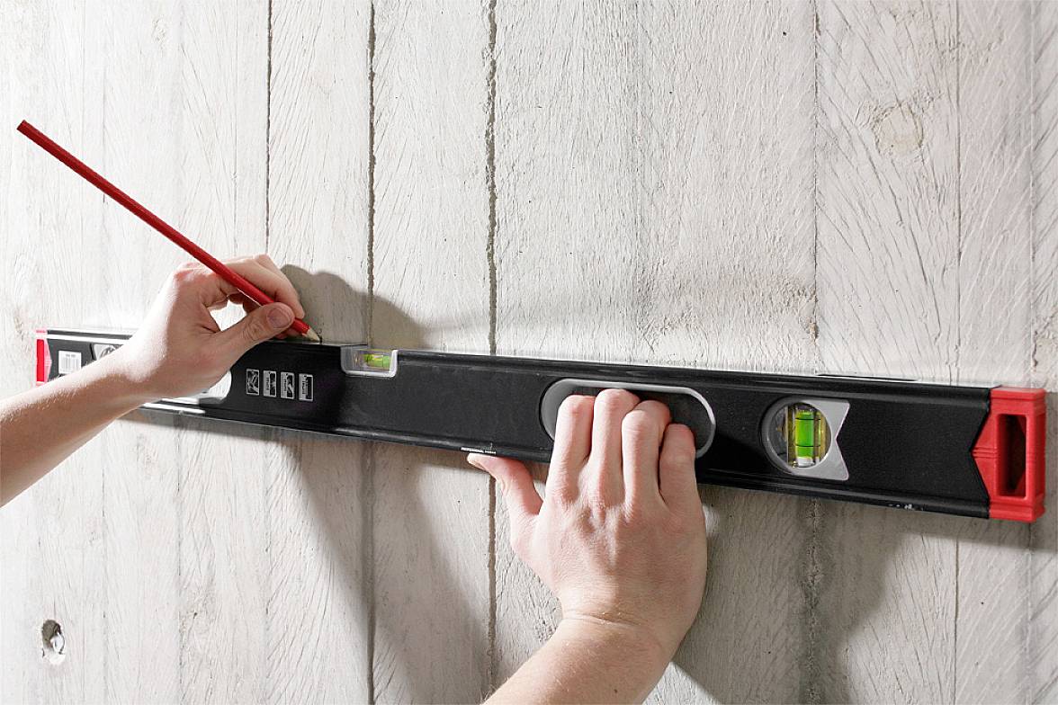 Eine Person markiert mit einem Bleistift eine Linie auf einer Holzwand, während sie eine Wasserwaage benutzt, um die Linie gerade auszurichten.