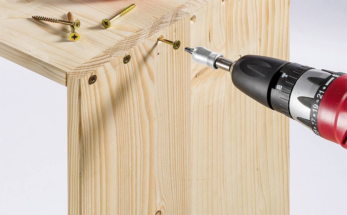 'Akkuschrauber zieht Schraube in Holzbalken ein, weitere Schrauben und Schraubenzieher in der Nähe, Nahaufnahme des Bauprozesses.'