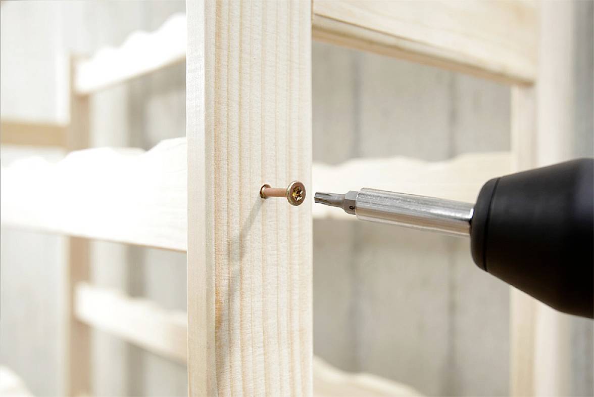 Eine Nahaufnahme eines Akkuschraubers, der eine Schraube in ein helles Holzbrett eindreht, zeigt den Aufbau eines Möbelstücks.