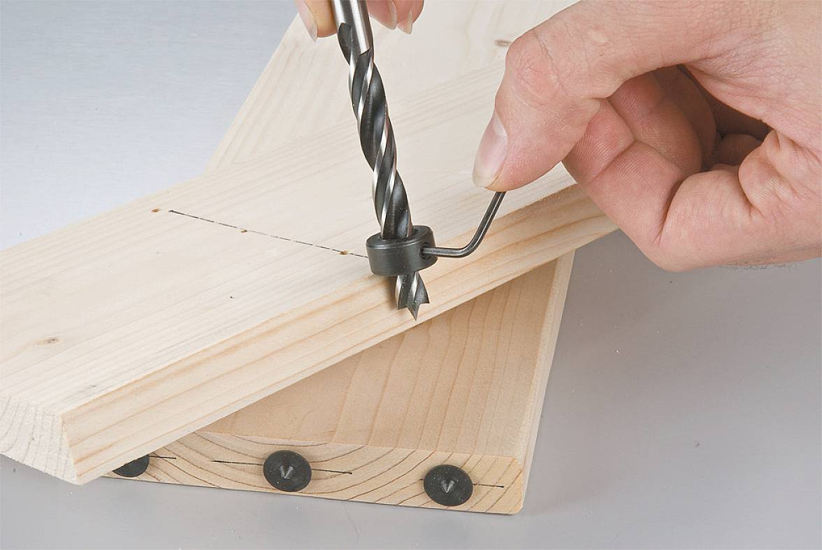 Holz wird mit einem Bohrer bearbeitet; ein Holzstück wird auf einem Ständer fixiert. Eine Hand hält den Bohrer.