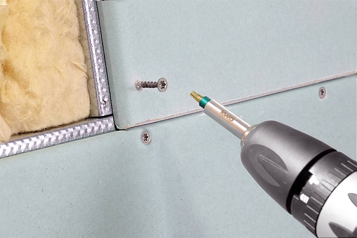 Bohrmaschine schraubt eine Schraube in eine Trockenbauwand ein. Links ist Dämmmaterial sichtbar. Hands-On-Werkzeug-Nutzung.