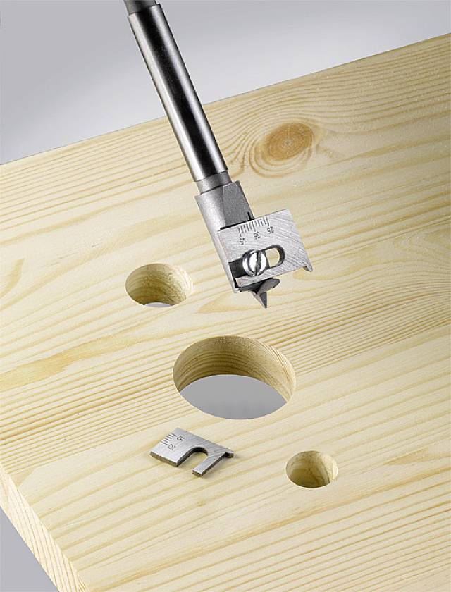 Holzbrett mit Bohrlöchern, ein Bohrkopf ist im Einsatz. Im Vordergrund liegt ein abgebrochener Teil des Bohrers auf dem Brett.