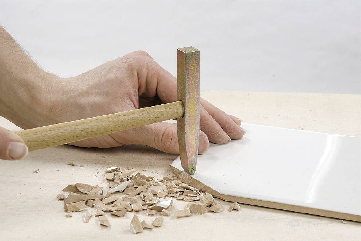 Eine Hand hält einen Hammer und zerschlägt eine Keramikkachel, wobei kleine Scherben entstehen.