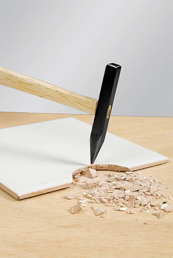 Ein Hammer bricht eine weiße Fliese auf einem Holztisch, wobei Fliesenscherben verstreut sind.