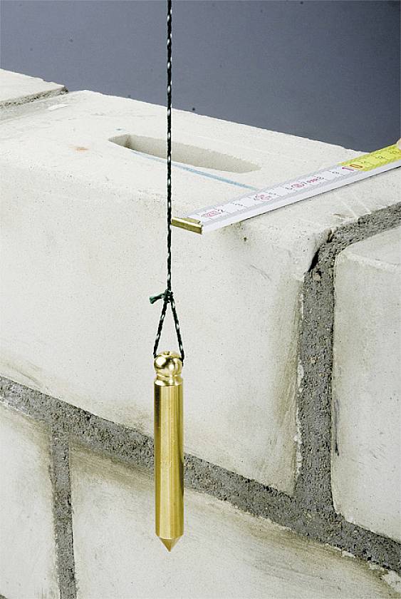 Ein Lot hängt an einer Schnur vor einer Mauer aus Betonblöcken. Ein Maßband liegt auf der Mauer, was auf Bauarbeiten hindeutet.