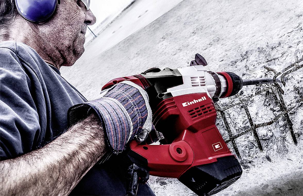 Eine Person benutzt einen roten Elektrohämmer, um in eine Betonoberfläche zu bohren. Die Person trägt Gehörschutz und Handschuhe.