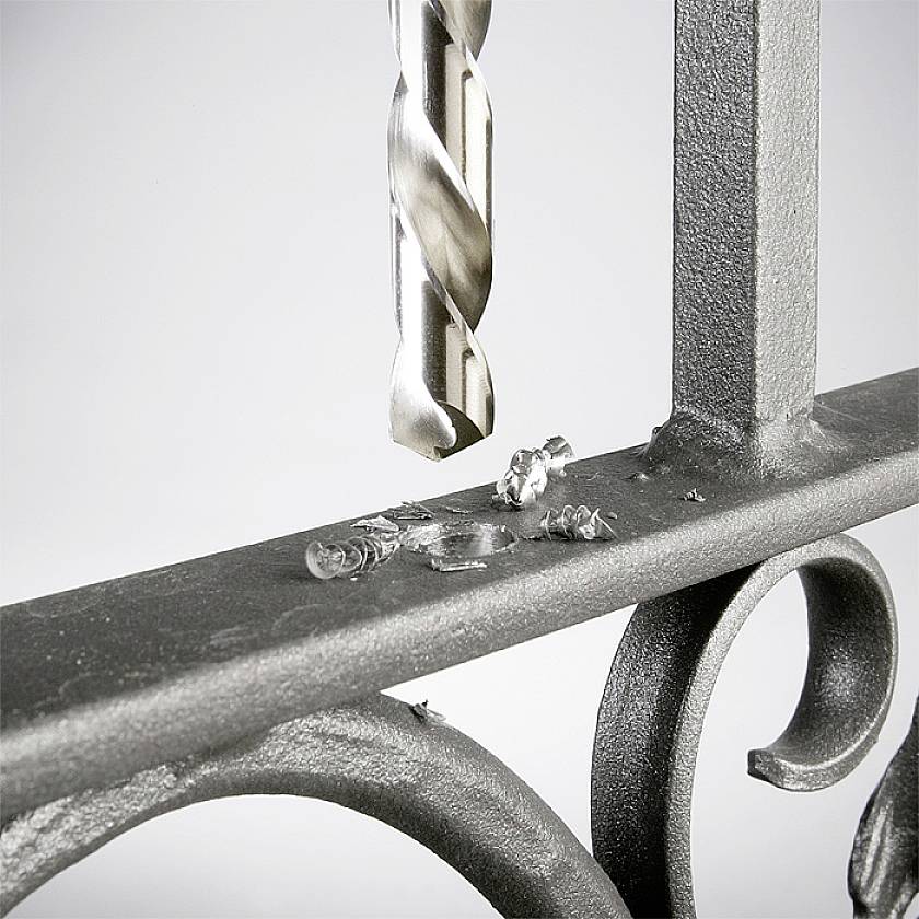 Metallbohrer durchdringt dickes Stahlblech, Späne fallen. Bild zeigt Präzision und Stärke beim Bohren, Fokus auf Gewindebohrer und Metall.