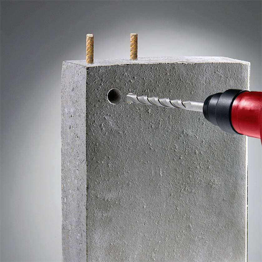 Eine rote Bohrmaschine bohrt ein Loch in einen grauen Betonblock mit zwei herausragenden Schrauben oben.