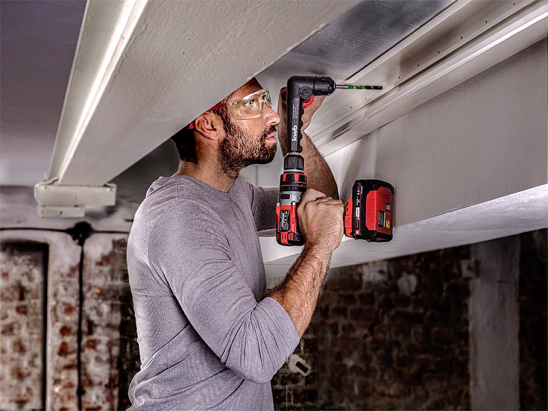 Ein Mann benutzt einen roten Akku-Bohrschrauber, um in eine weiße Decke zu bohren. Er trägt eine Schutzbrille und befindet sich in einem Keller.