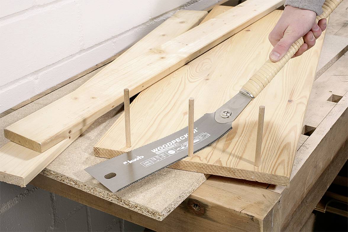 Eine Hand sägt mit einer Japansäge Holzleisten auf einer Werkbank. Drei Holzleisten stehen senkrecht auf einem Brett aus hellem Holz.