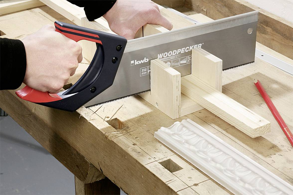 Eine Person schneidet ein Stück Holz mit einer Handsäge auf einer Werkbank. Es sind weitere Holzstücke und ein Bleistift zu sehen.