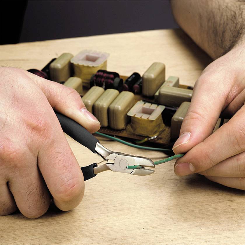 Eine Person schneidet mit einer Zange ein grünes Kabel. Im Hintergrund sind elektronische Bauteile auf einer Platte zu sehen.