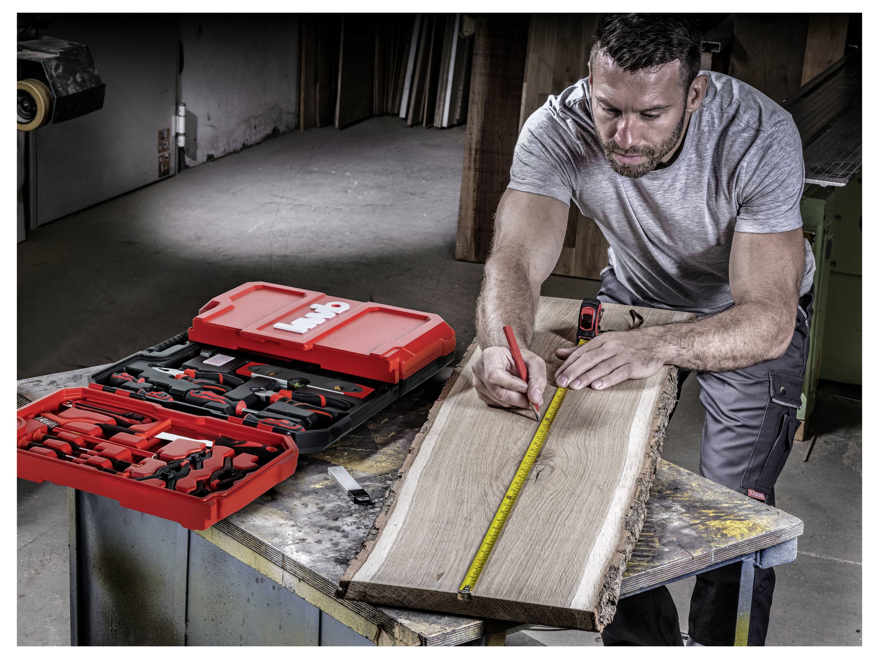 Ein Mann misst eine Holzplanke mit einem Maßband auf einem Werkstatttisch. Werkzeugkästen mit roten Gehäusen sind neben ihm geöffnet.