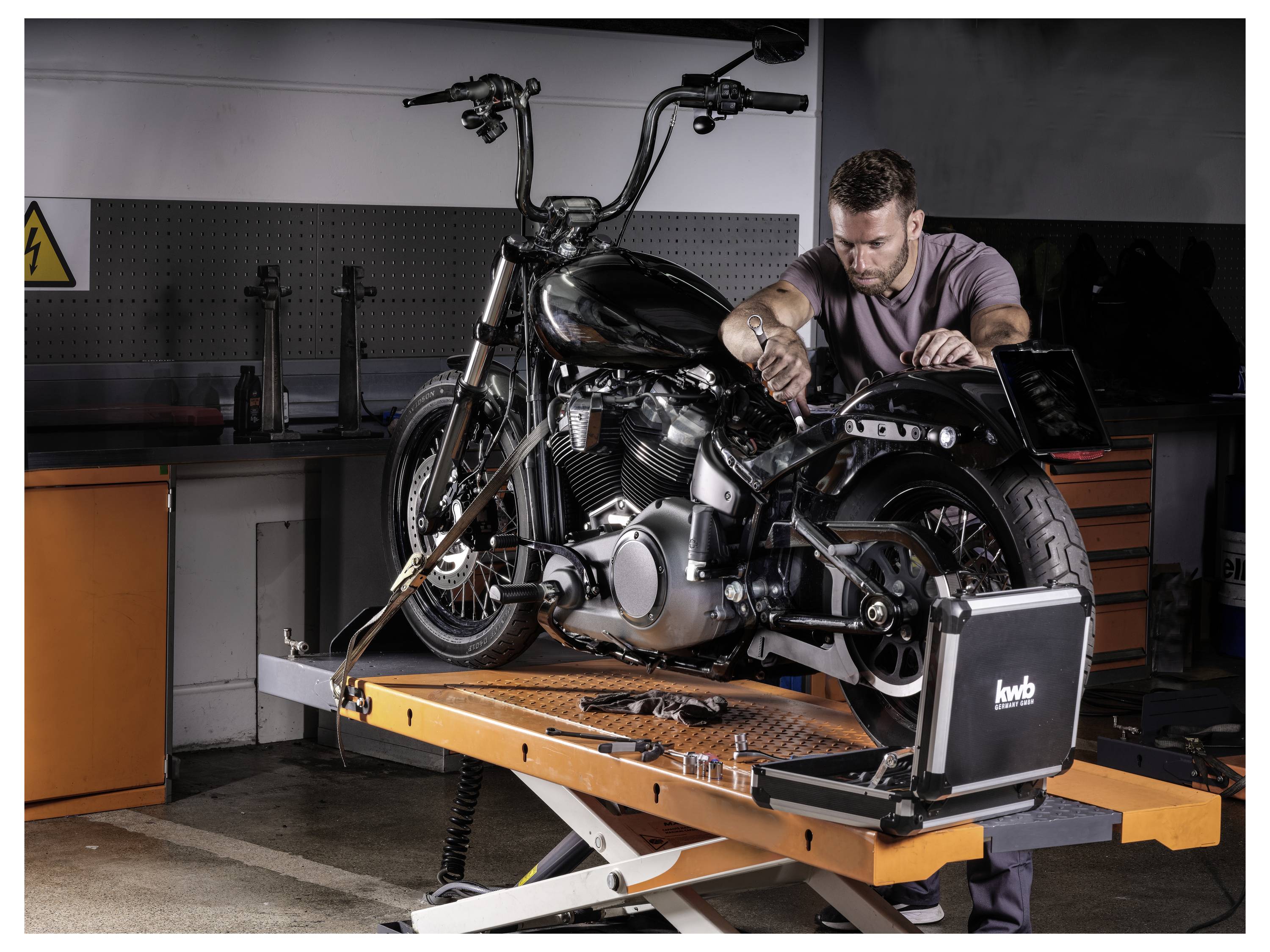 Eine Person repariert ein Motorrad auf einer Hebebühne in einer Garage und konzentriert sich auf den Motorbereich. Werkzeuge und Ausrüstung sind im Hintergrund sichtbar.