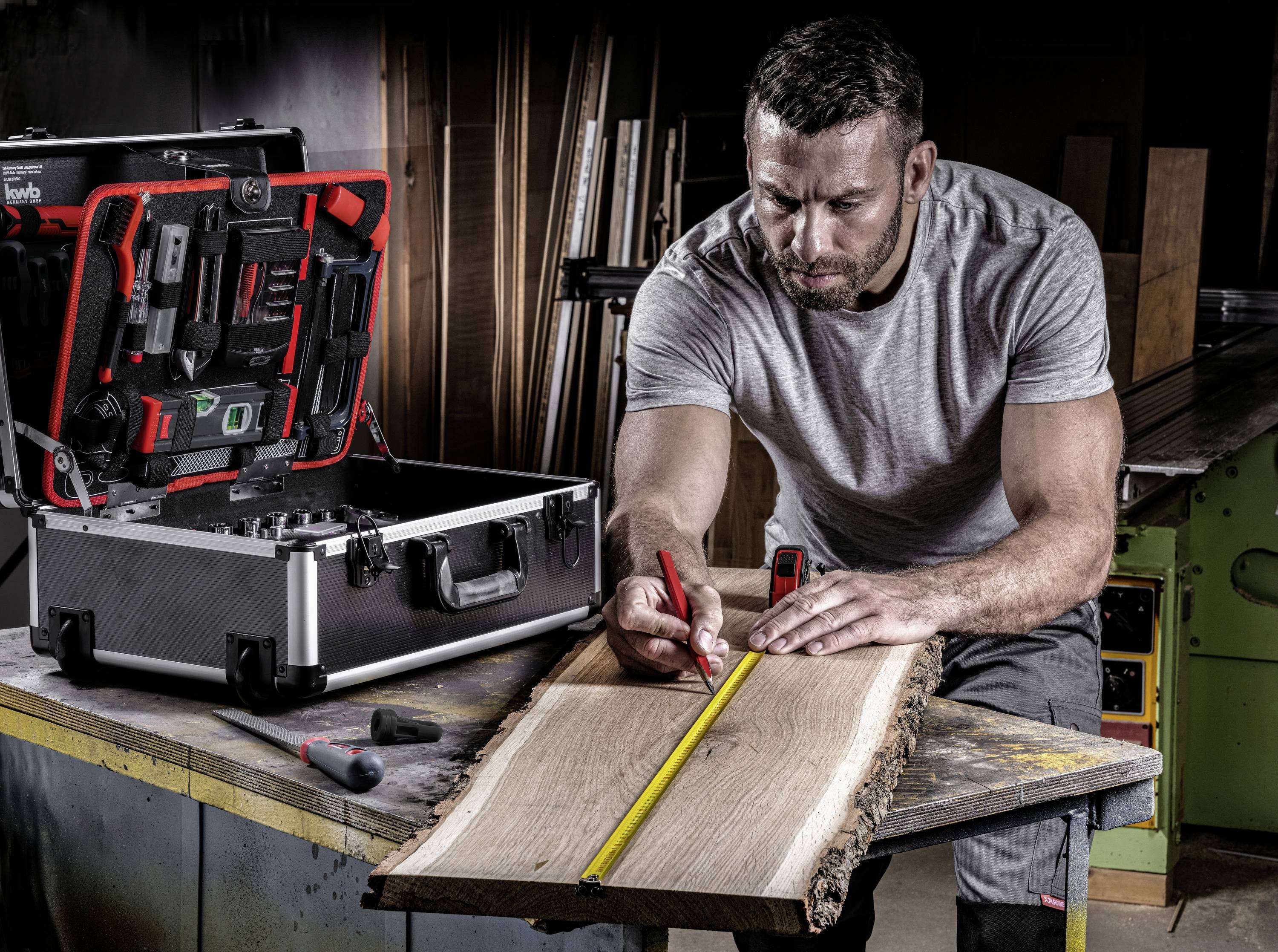 Ein Mann misst ein Stück Holz auf einer Werkbank in einer Werkstatt. Neben ihm steht ein offener Werkzeugkoffer mit verschiedenen Werkzeugen.