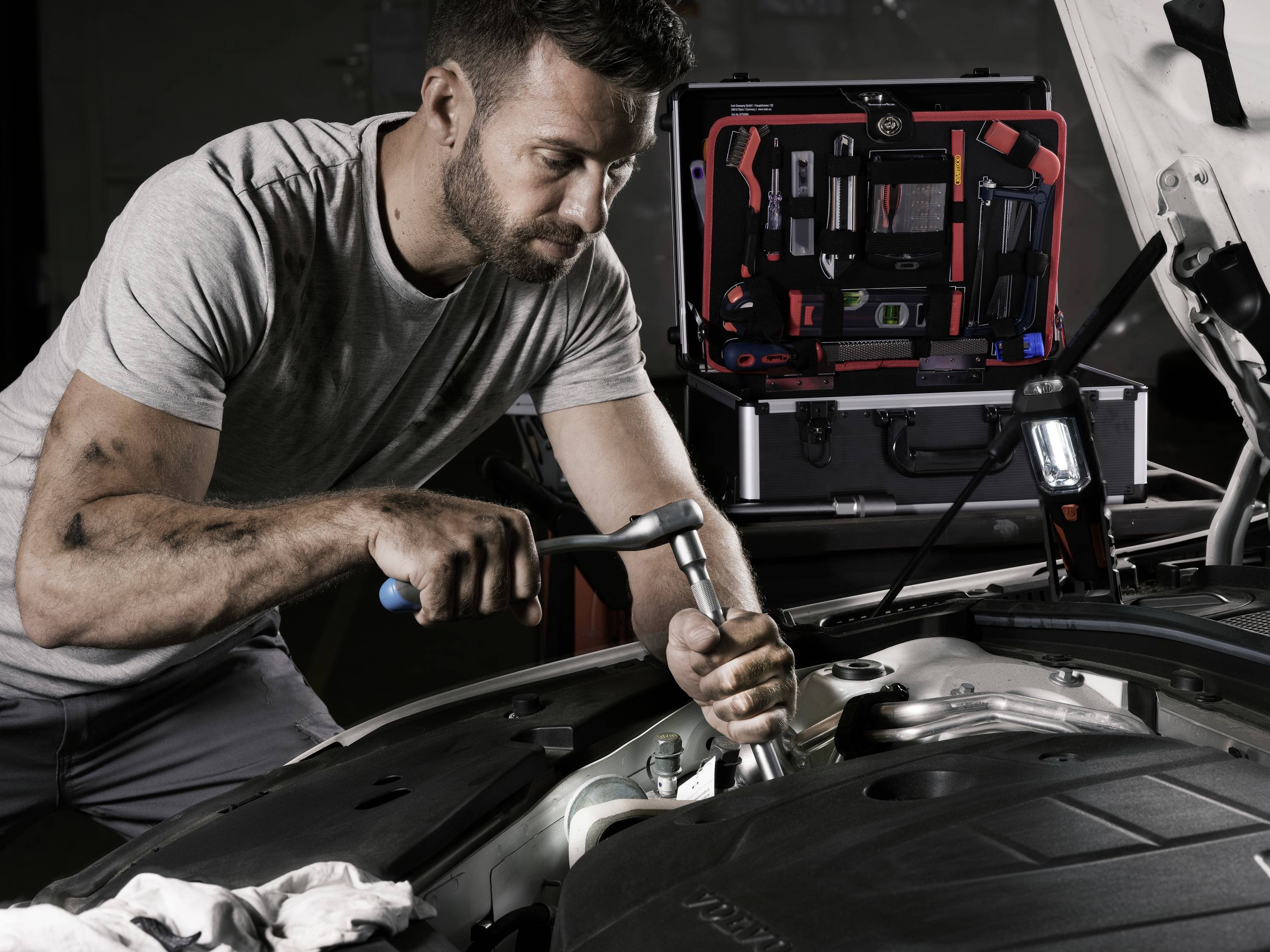 Mechaniker repariert Auto im Motorraum. Werkzeuge sind um ihn herum, er konzentriert sich auf eine Schraube. Sein Gesicht zeigt Entschlossenheit.