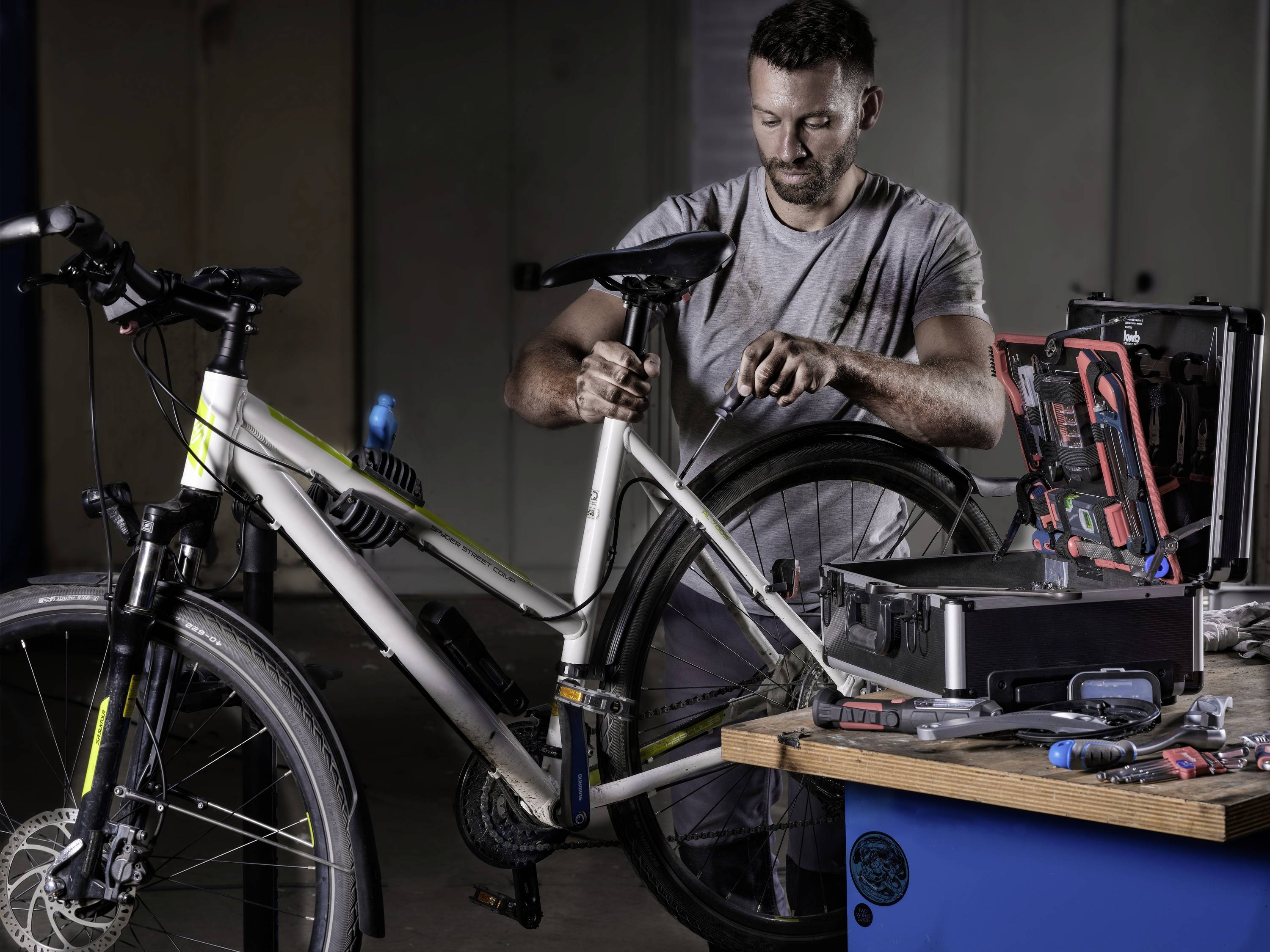 Ein Mann arbeitet in einer Werkstatt an einem Fahrrad, umgeben von Werkzeugen und Ausrüstung auf einem Tisch.
