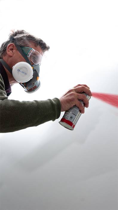 Eine Person trägt eine Schutzmaske und sprüht rote Farbe aus einer Sprühdose gegen eine weiße Wand.