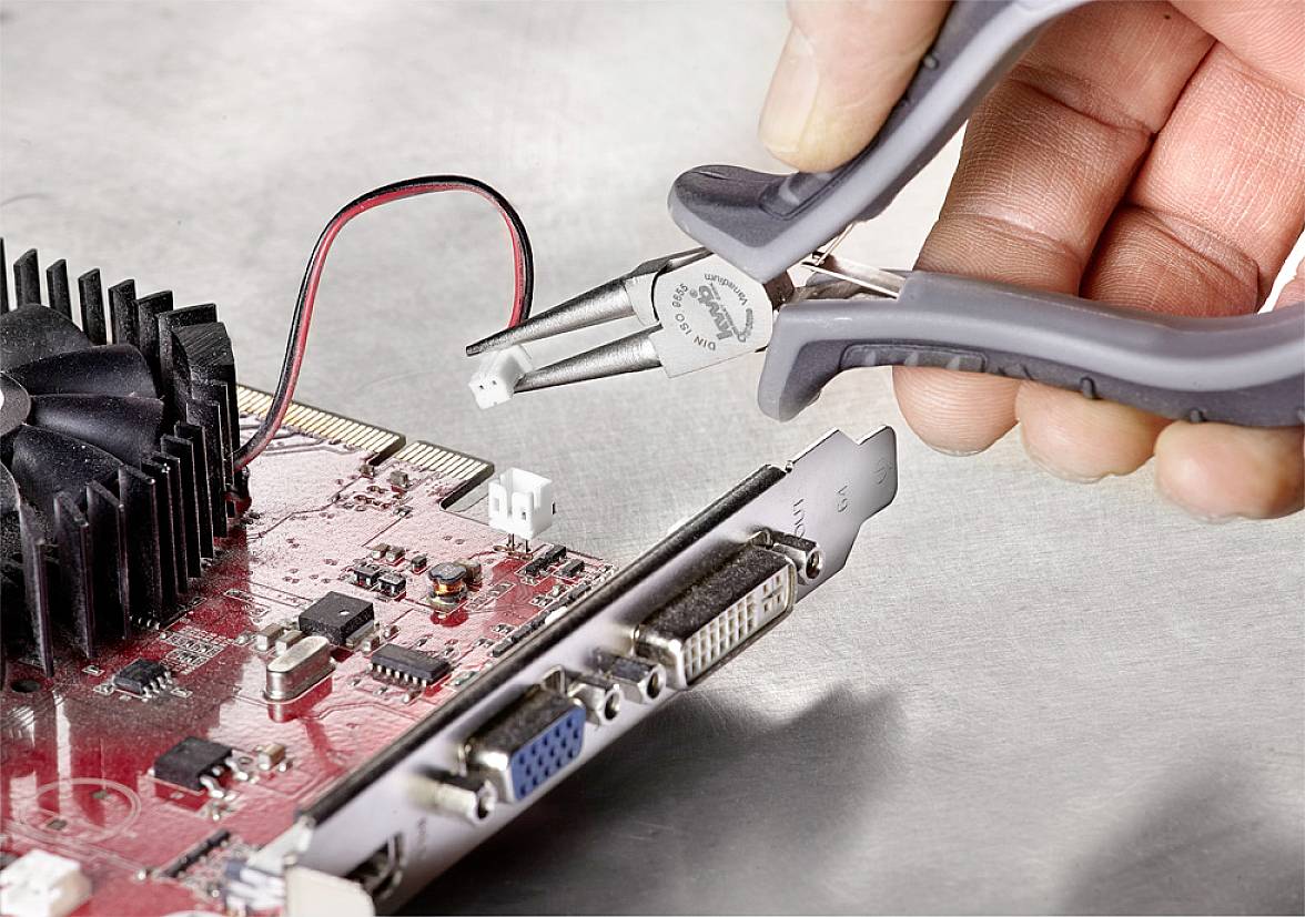 Eine Hand hält eine Zange und arbeitet an einer roten Computerplatine mit sichtbaren Anschlüssen und einem Kühlkörper.