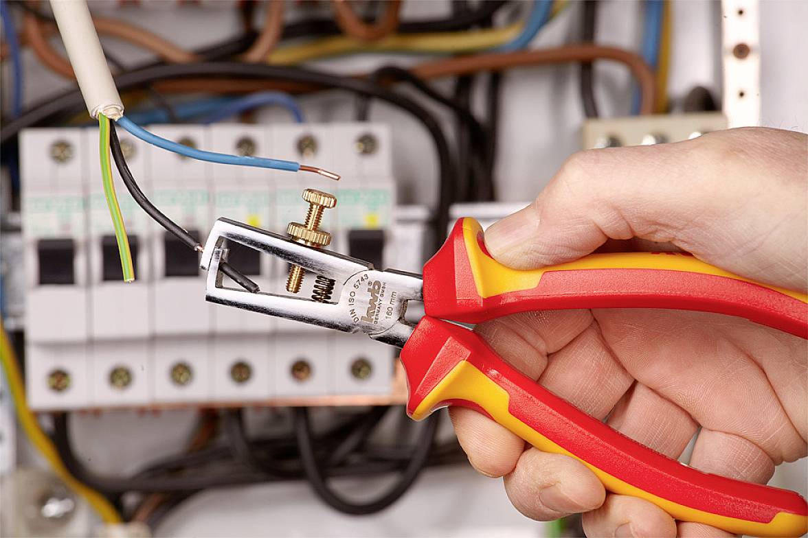 Eine Hand hält einen Abisolierer, um Kabel in einem Schaltschrank freizulegen. Elektrische Komponenten im Hintergrund.