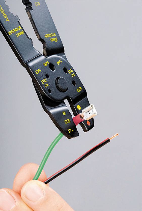 Eine Hand hält ein grünes und ein rotes Kabel, während eine Crimpzange einen Kabelschuh am roten Kabel befestigt.